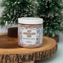 Load image into Gallery viewer, A container of HOT CHOCOLATE BAR WHIPPED SOAP by Bath Bevy, placed on a wooden surface with a decorative pattern and Christmas tree branches in the background.