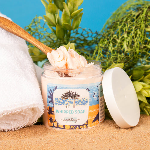 Jar of 'Beach Bum Whipped Soap' with a wooden spoon on sand, towel, and plants in the background.