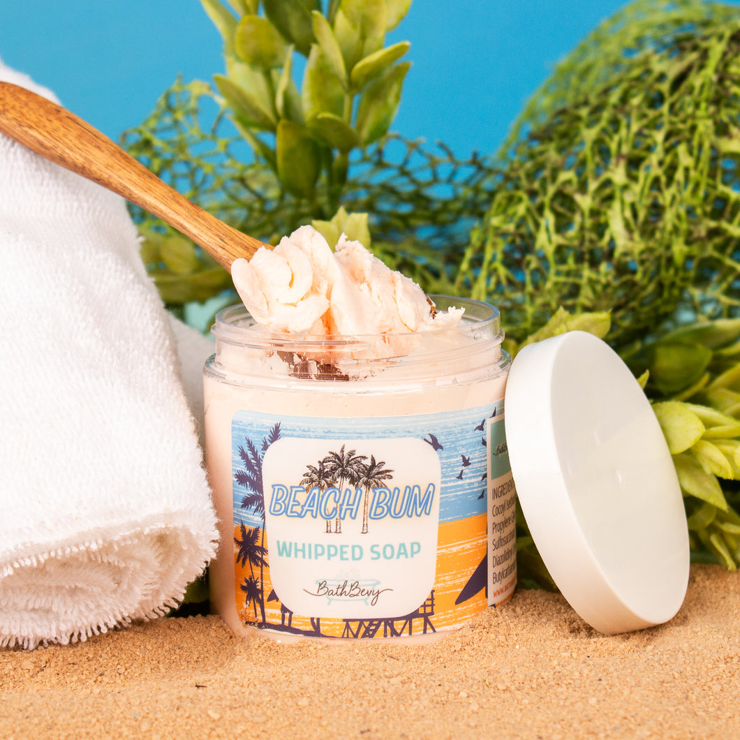 Jar of 'Beach Bum Whipped Soap' with a wooden spoon on sand, towel, and plants in the background.