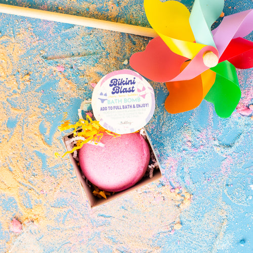 Pink bath bomb in a box with a colorful pinwheel on a textured blue and gold background
