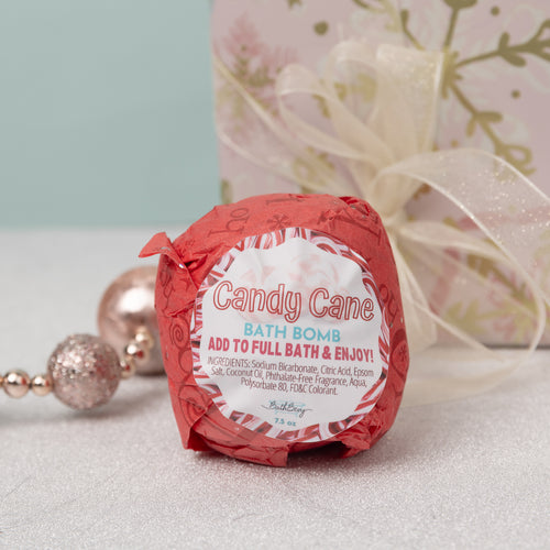 A red bath bomb labeled 'Candy Cane' with a Christmas-themed decoration on a light background, accompanied by a gift ribbon and decorative balls.