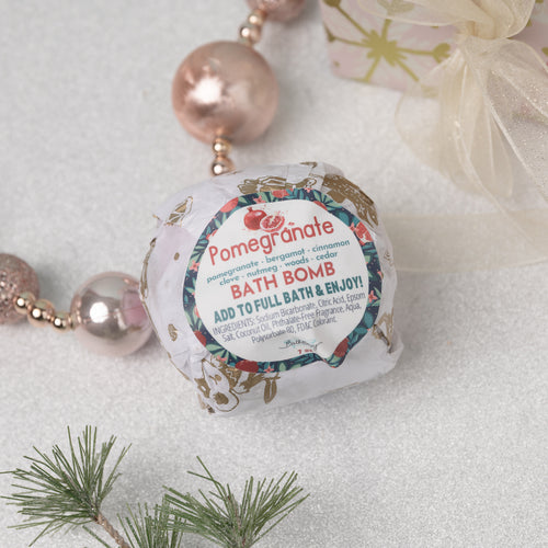 A packaged bath bomb with pomegranate and spice botanical print design, labeled 'Pomegranate Bath Bomb', placed on a decorative setting with pearls and greenery.