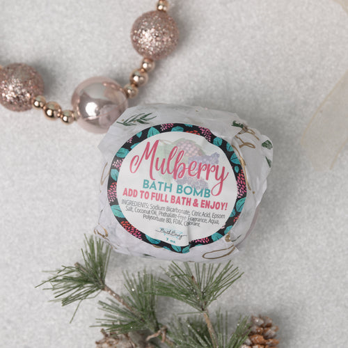 A packaged bath bomb with 'MULBERRY BATH BOMB' label, placed on a surface with pine branches and decorative balls, indicating it is meant to add to a full bath and enjoy the fruit-themed scent and therapeutic benefits.
