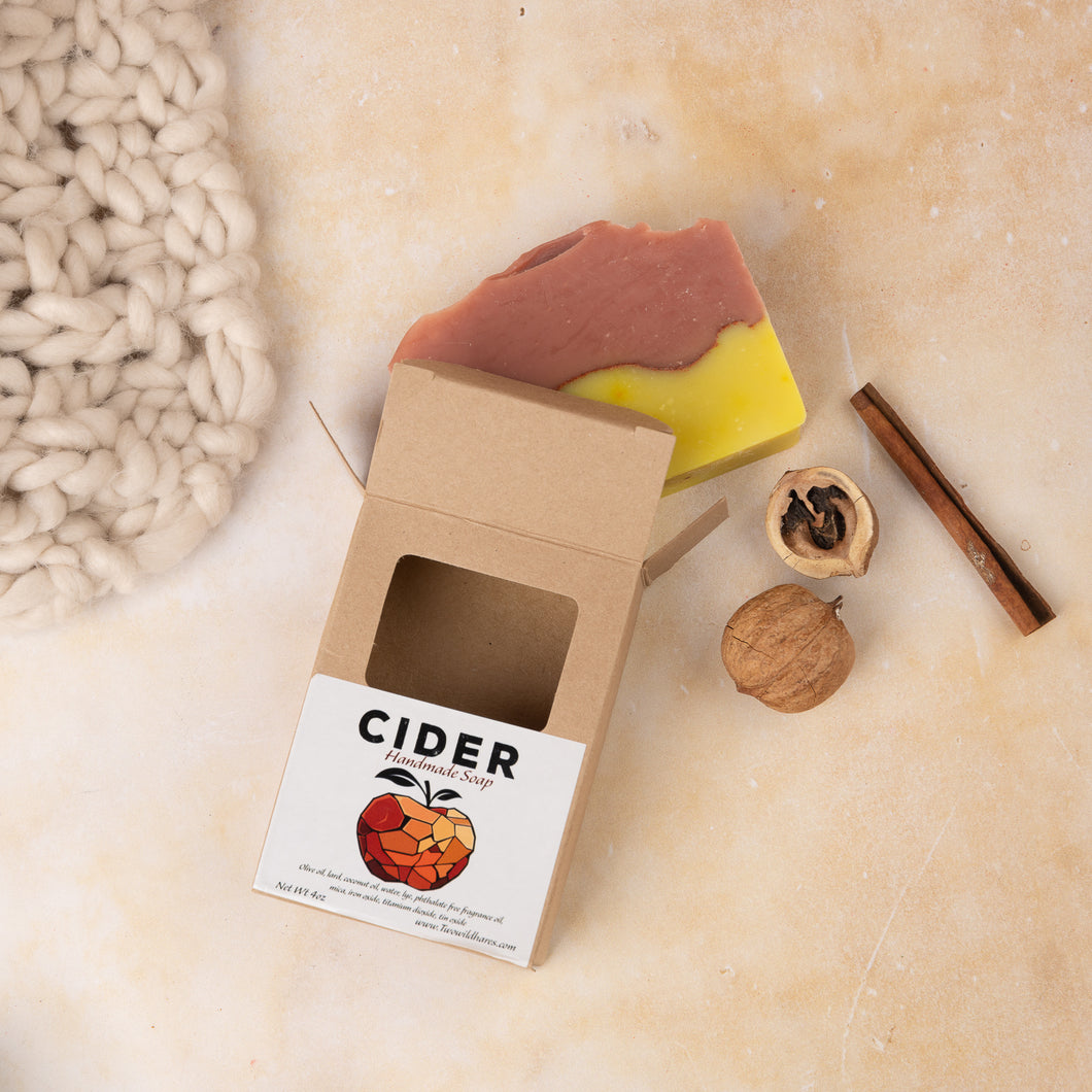 A handcrafted soap bar with a piece of cider apple, a cinnamon stick, and some dried leaves, all placed on a marble surface next to a knitted fabric.