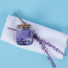Load image into Gallery viewer, Purple candle in a glass jar with a wooden lid on a white towel against a blue background