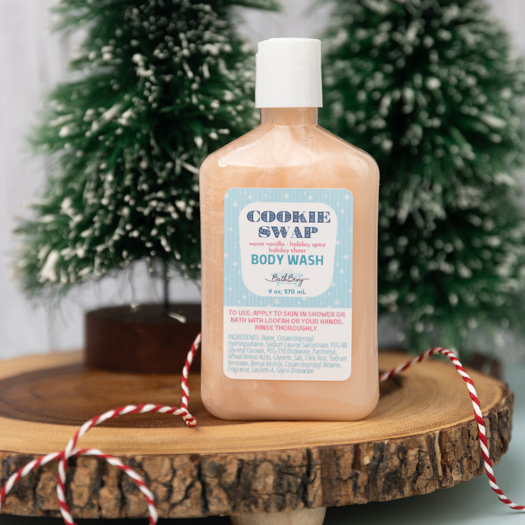 A bottle of Cookie Swap body wash by Burt's Bees, pump bottle design, with a label that reads 'holiday'. The bottle is placed on a wooden surface with a decorative backdrop of Christmas trees and red and white twine.