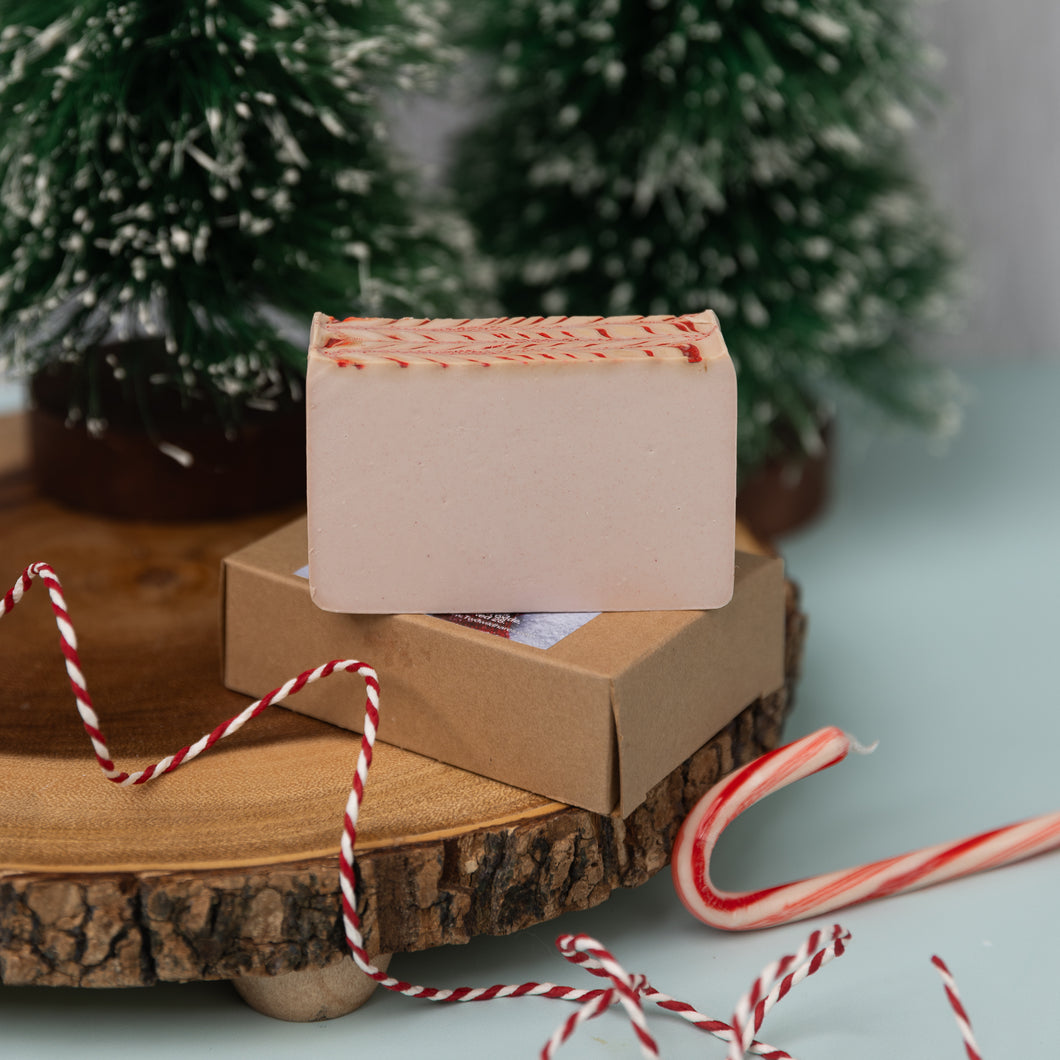 A handmade soap bar with red and white striped candy cane decorations on a wooden surface, surrounded by holiday-themed decorations including a artificial Christmas tree and candy canes.