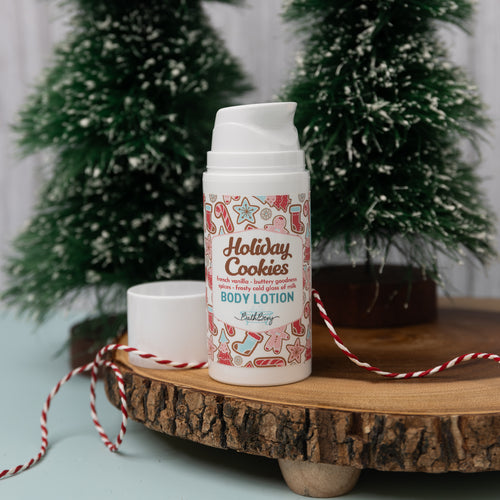 A bottle of HOLIDAY COOKIES BODY LOTION by Bella Dahl, placed on a wooden surface with a decorative background featuring Christmas trees and ornaments.