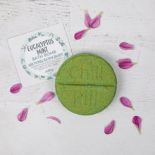 Load image into Gallery viewer, Green bath bomb with eucalyptus mint label on a light wooden surface with purple flowers.